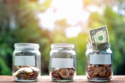 Three glass jars filled with coins and a dollar bill, representing small donations adding up
