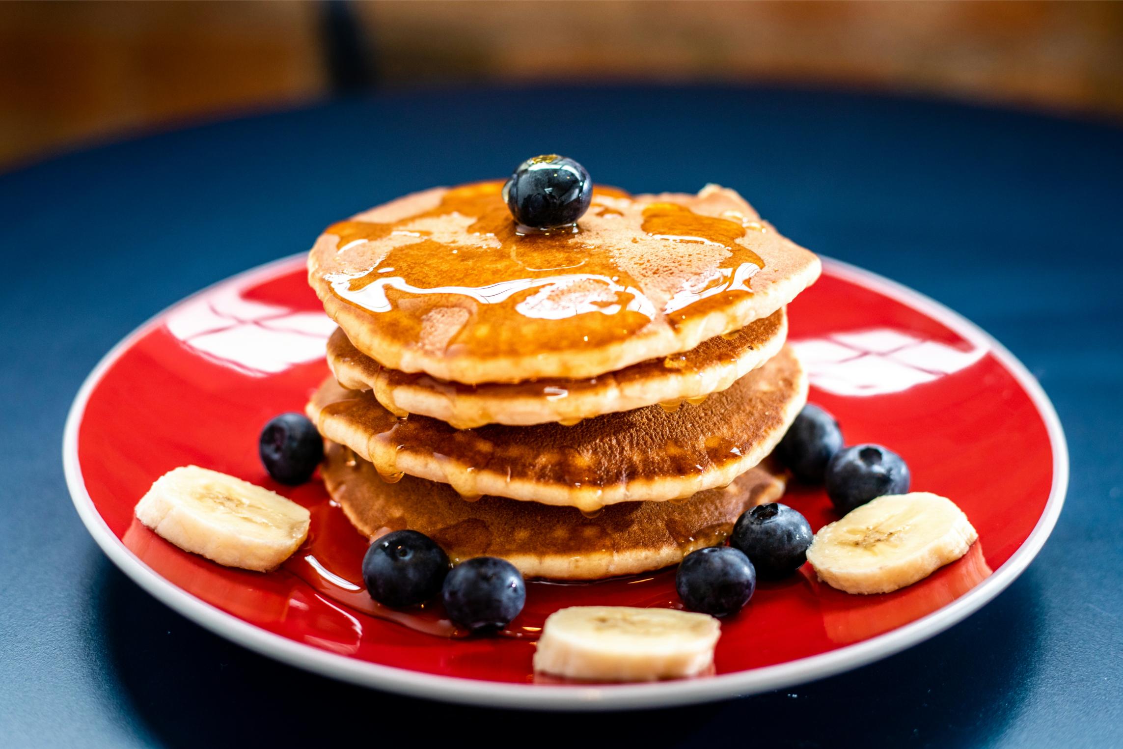 pancake-breakfast-fundraiser A stack of pancakes drizzled with syrup, topped with blueberries and banana slices on a red plate — pancake breakfasts are a classic sales fundraiser.